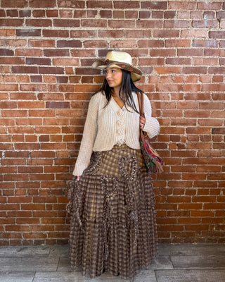 A woman stands against a red-brown brick wall wearing a cream ribbed cardigan, a brown plaid maxi skirt with floral applique, a beige cowboy hat, and carrying a patterned shoulder bag.