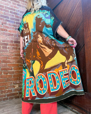 A woman stands in a corner wearing a long, loose black tunic-style top with a large colorful western rodeo graphic. The print shows a cowboy on a horse and the word 'RODEO' in large marquee-style letters.