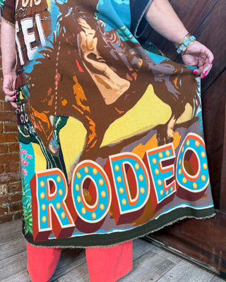 A person holds up a colorful woven blanket featuring a cowboy riding a horse and the word 'RODEO' in large marquee-style letters. The blanket has a multicolor design with brown, yellow, blue, orange, and red tones, and the person is wearing coral-pink pants.