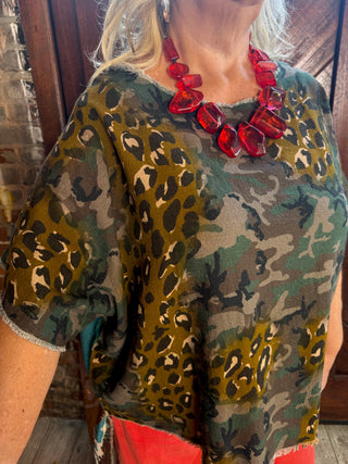 A close-up of a woman wearing a short-sleeve camouflage and leopard print top with a red chunky statement necklace, standing in front of a rustic wooden door.
