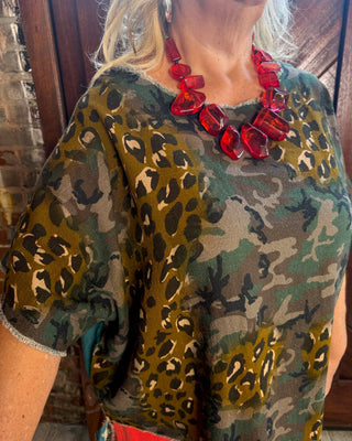 A close-up of a woman wearing a short-sleeve camouflage and leopard print top with a red chunky statement necklace, standing in front of a rustic wooden door.
