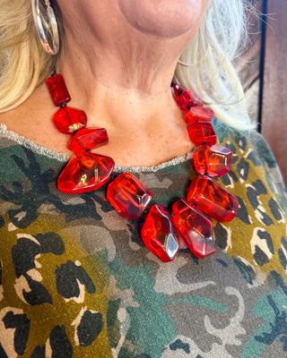 A close-up of a woman wearing a chunky statement necklace made of faceted translucent red beads, paired with a green and black leopard print top.