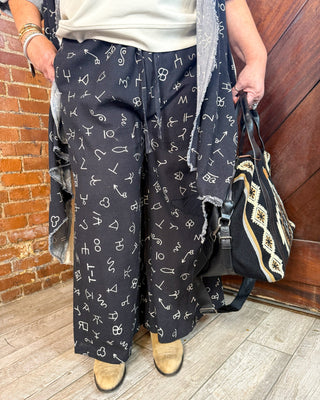 A person wearing black wide-leg pants covered in a white script and symbol print, styled with a cream top, a gray cardigan, tan boots, and carrying a black and cream patterned handbag.