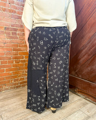 A person wearing black wide-leg pants with a white abstract symbol print, paired with a cream-colored top, standing in a room with a brick wall and wooden floor.