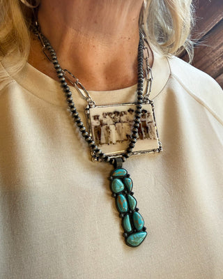 A close-up of a person wearing a layered necklace. The necklace features a dark beaded chain with a rectangular pendant showing a sepia-toned family photograph, layered over a silver chain with a vertical drop of turquoise-colored stones on a black cord.