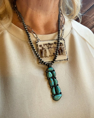 A close-up of a person wearing a layered necklace. The necklace features a dark beaded chain with a rectangular pendant showing a sepia-toned family photograph, layered over a silver chain with a vertical drop of turquoise-colored stones on a black cord.