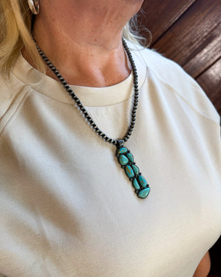 A close-up of a person wearing a black beaded necklace with a turquoise stone pendant, paired with a cream-colored top.