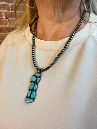 A close-up of a person wearing a long beaded necklace with a vertical pendant featuring bright turquoise-blue stones set in dark metal, paired with a cream sweatshirt and a brick wall background.