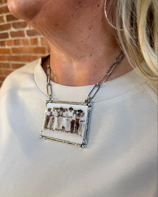 A close-up of a person wearing a rectangular pendant necklace with a vintage-style photograph of people in white clothing, set in a silver-toned metal frame and hanging from a silver chain.