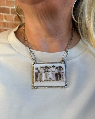 A close-up of a person wearing a rectangular silver pendant necklace with a vintage-style black and white photograph of a group of people, attached to a silver chain.