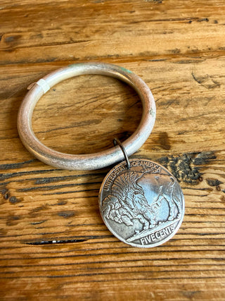A rustic silver-toned metal bangle bracelet with a dangling coin charm, displayed on a textured wooden surface.
