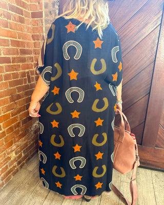 A woman viewed from behind wearing a loose black maxi dress with an all-over pattern of orange stars, olive green horseshoes, and white horseshoes. She is standing in a rustic indoor setting with a brick wall, wooden door, and light wood floor, holding a tan shoulder bag.