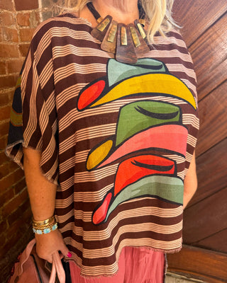 A woman wearing a brown and beige horizontally striped tunic top with a large, colorful abstract graphic print on the front. The print features wavy shapes in yellow, green, pink, and red.