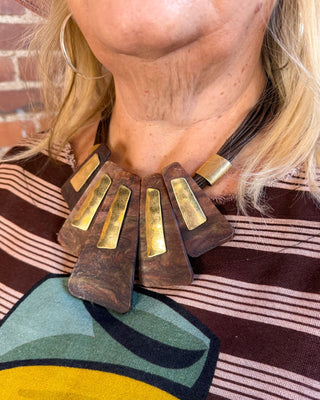 A close-up of a person wearing a chunky statement necklace with dark brown wooden disc elements and shiny gold-toned metal bars, paired with a brown and pink striped shirt.
