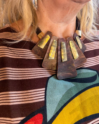 A close-up of a woman wearing a chunky statement necklace made of dark brown fan-shaped pieces with gold-toned rectangular inlays, paired with a brown and beige striped top.