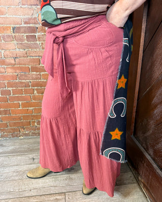 A person wearing dusty rose wide-leg tiered pants with a side tie and a dark navy patterned fabric panel on the side, standing on a wood floor against a brick wall.