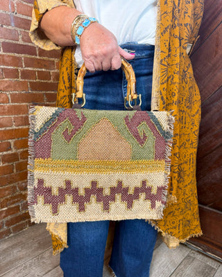 A person holding a woven rectangular handbag with two tan handles. The bag has a geometric Southwestern-style pattern in beige, olive green, mustard yellow, and brown.