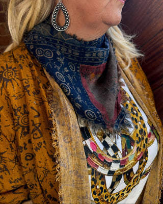 A close-up side profile of a woman wearing a layered fashion look featuring a blue patterned scarf, a mustard yellow patterned cardigan, and a white graphic t-shirt with a leopard print and gold chain necklace.