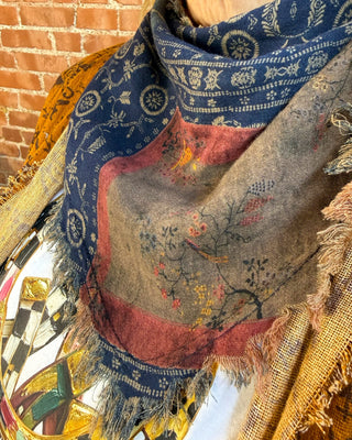 A multicolor patterned scarf draped on a mannequin, featuring a navy blue border with cream motifs, a central gray-brown panel with a bird and floral design, and rust-red borders, set against a brick wall background.