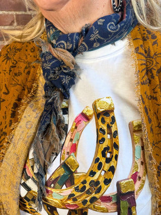 A close-up of a person wearing a white graphic t-shirt with a large gold horseshoe design, styled with a blue paisley scarf and a mustard yellow patterned cardigan.