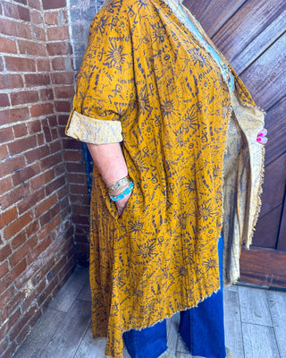 A person wearing a mustard yellow tunic-style top with a black printed pattern, blue pants, and a beige shawl, standing in a corner with a brick wall on one side and a wooden door on the other.