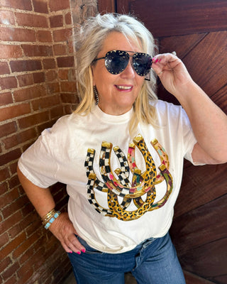 A woman wearing a white graphic t-shirt with a large snakes and checkerboard print, blue jeans, and black sunglasses, posing in front of a brick wall and wooden door.