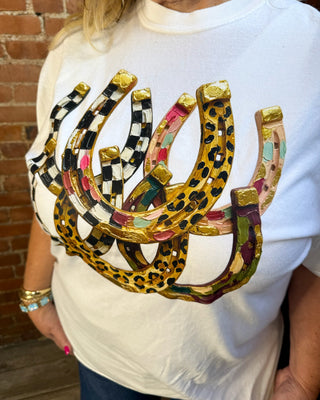 A white crewneck t-shirt featuring a large graphic print of six overlapping horseshoes in various patterns (leopard print, checkered black and white, pink, green, and gold), worn by a model standing in front of a brick wall.
