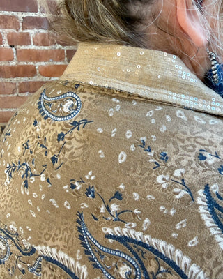 Close-up of a person wearing a tan and navy paisley patterned jacket with sequined detailing on the collar, standing in front of a brick wall.