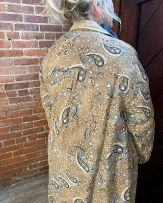 A person viewed from behind wearing a tan paisley jacket with blue and white patterns, standing in front of a red brick wall and dark wooden door.