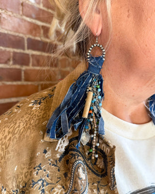 A close-up of a person wearing a long, bohemian-style dangle earring made from blue denim ribbon, beads, and a small wooden bead, shown against a brick wall background.