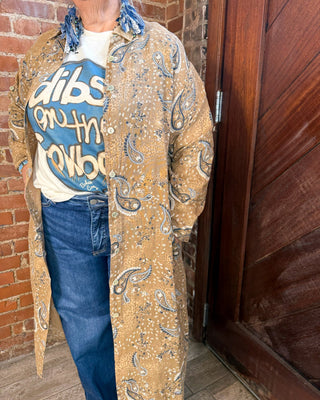A person wearing a tan paisley bandana-print longline cardigan over a graphic t-shirt and blue jeans, standing in front of a brick wall and a dark wooden door.