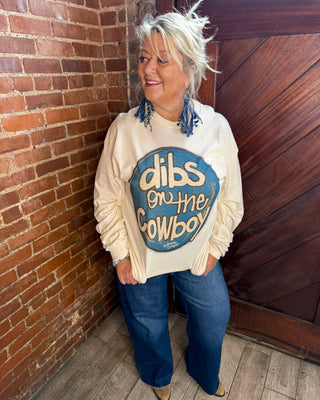 A woman with platinum blonde hair stands in a brick and wood doorway wearing a cream-colored sweatshirt with a blue graphic that reads 'dibs on the Cowboy' and blue jeans.