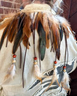 A close-up of a person wearing a decorative feathered garment or accessory with long brown, tan, and dark feathers, beaded cords, and tassels over a distressed off-white top. A brick wall is visible in the background.