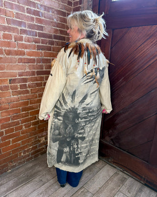 A woman stands in a corner with a brick wall and dark wooden door, wearing a long cream-colored distressed jacket with Native American-inspired feather and bead trim and black graphic print on the back.
