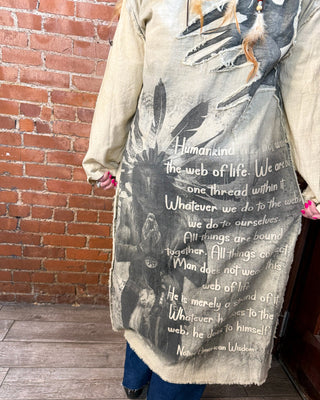 A person wearing a long beige tunic-style top with a large black and gray graphic print featuring a Native American headdress, feathers, and text, standing in front of a red brick wall.
