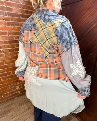 A person shown from the back wearing a patchwork quilted shirt jacket with star appliqués, standing in a room with a brick wall and wooden door.