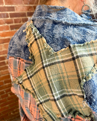 A close-up of a person wearing a handmade patchwork shirt with multiple fabric panels, including a prominent green and tan plaid patch, a blue floral patch, and a blue denim patch, set against a brick wall background.