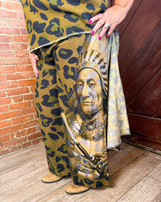 A person wearing wide-leg pants with an olive green and black leopard print base and a large front graphic of a Native American portrait, standing on a wood floor between a brick wall and a dark wooden door.