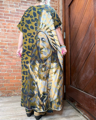 A woman viewed from behind wearing a loose, short-sleeve maxi dress with a leopard-print background and a large Native American portrait printed on the back. She stands between a red brick wall and a dark wooden door.