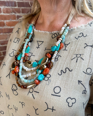 A close-up of a woman wearing a multi-strand statement necklace with turquoise, orange, white, and brown beads over a beige zodiac-sign printed top, set against a brick wall background.