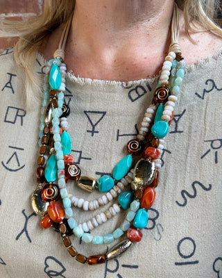 A close-up of a person wearing a multi-strand statement necklace with turquoise, orange, brown, and white beads over a beige patterned top.