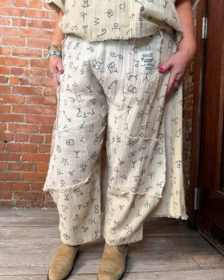 A person wearing loose, frayed beige pants covered in a black symbol print, standing indoors against a brick wall and wooden door. The top is partially visible and tan suede boots are worn with the pants.