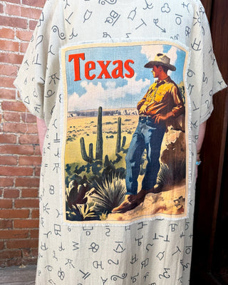 A light beige short-sleeve tunic or shirt with a black alphabet print, featuring a large square graphic on the back depicting a cowboy in a desert landscape with the word 'Texas' in red text.