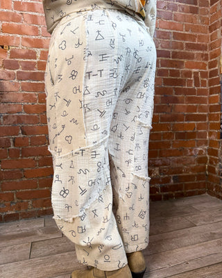 A person wearing loose, cream-colored patchwork pants covered in black handwritten symbols, standing in a room with a red brick wall and wood floor.