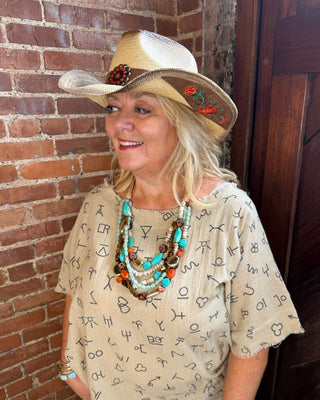 A woman wearing a beige patterned short-sleeve top, matching pants, a straw cowboy hat with red floral embroidery, and layered turquoise and brown beaded necklaces, standing in front of a brick wall and dark wooden door.