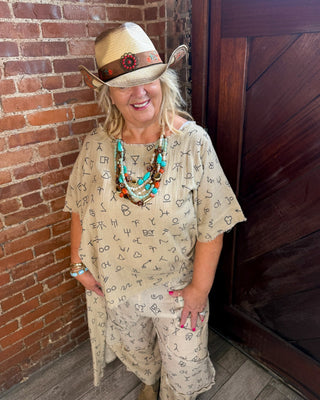 A woman wearing a beige bohemian-style outfit with a matching hat, layered turquoise and brown bead necklaces, and bracelets, standing in a rustic room with a brick wall and dark wooden door.