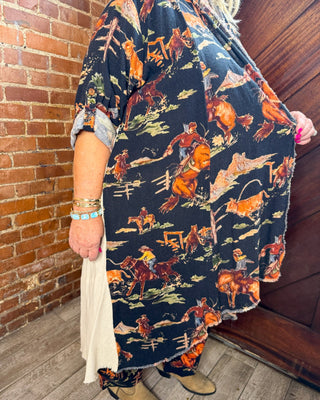 A person wearing a loose black tunic-style top with a colorful cowboy and horse print, paired with tan boots, standing indoors against a brick wall and wooden door.