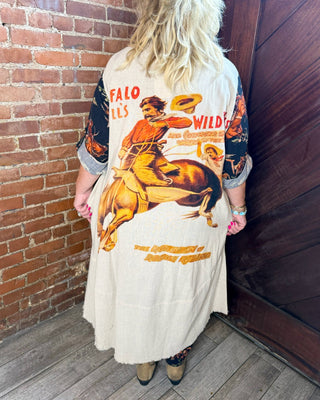 A woman stands with her back to the camera in a rustic indoor corner with a brick wall and dark wooden door, wearing a long cream-colored cardigan or dress with a large vintage western buffalo rider graphic on the back and patterned sleeves.