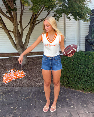 A blonde woman stands outdoors on a paved walkway wearing a white and orange Tennessee cheerleader crop top, denim shorts, and white sandals. She holds an orange and white pom-pom in one hand and a football in the other.