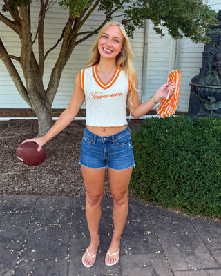 A smiling blonde woman stands outdoors on a paved walkway wearing a white Tennessee cheerleader-style crop top with orange trim, blue denim shorts, and white sandals. She holds a football in one hand and orange-and-white pom-poms in the other, with a tree, shrubs, and a white building in the background.